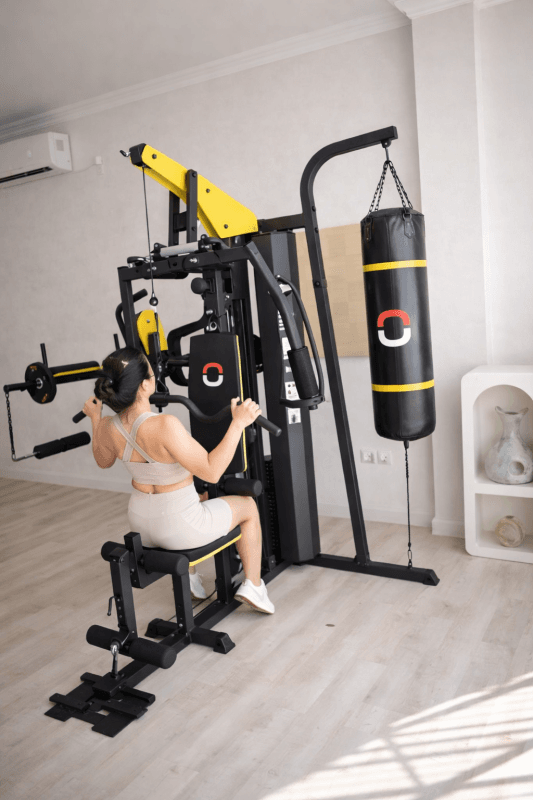 Wanita berolahraga menggunakan mesin home gym hitam kuning dengan punching bag di ruangan terang.